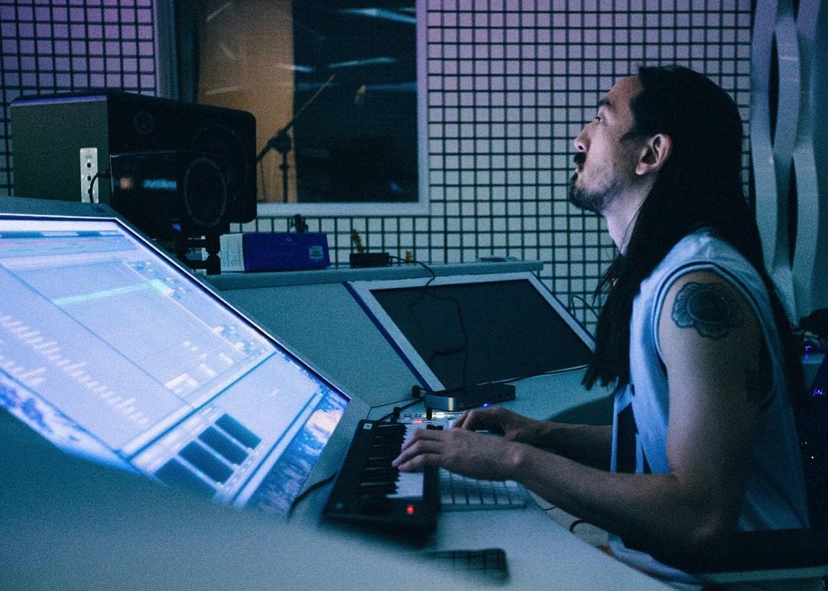 Steve Aoki playing on his keyboard while facing straight ahead, in the process of producing new music from his dimly-lit studio, the Neon Future Cave