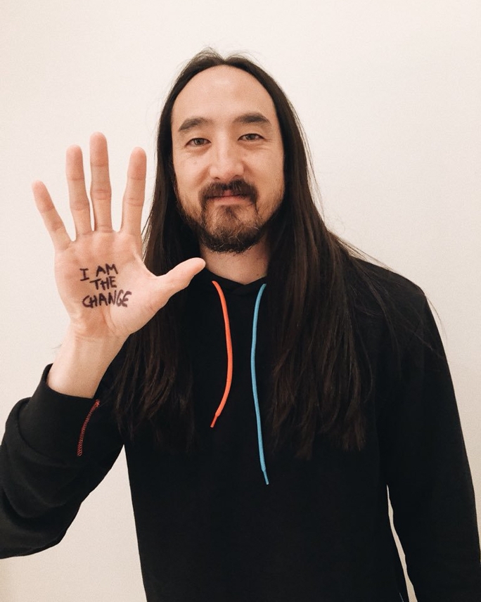 A headshot of Steve Aoki holding up his hand with the words "I am the change" written on his palm