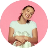 A studio PR photo portrait of Millie Bobby Brown smiling with her head leaned to the side, hand laid over the other, against a pink backdrop