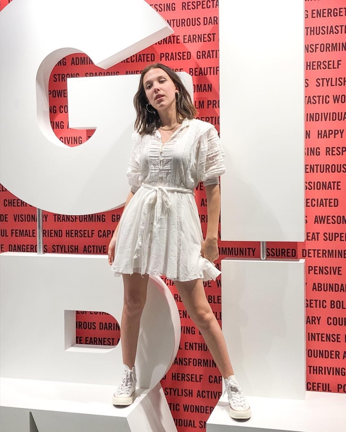 Millie Bobby Brown standing confidently in a white dress and sneakers in front of big blocked letters spelling out "GIRL" in white against a pink wall