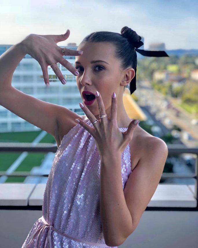 Millie Bobby Brown showing off her freshly manicured nails as she sports a pink sequined dress against a cityscape photography backdrop