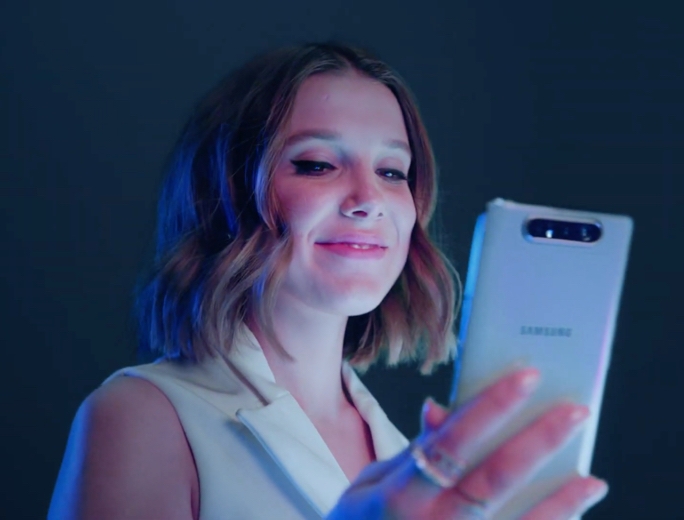 Millie Bobby Brown smiles as she looks at a prism white Galaxy S10 in her hand