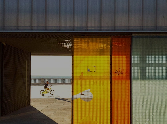 A little boy rides his bicycle on a boardwalk on a sunny day, the ocean as his backdrop. The view of him is captured inside an open building behind yellow, red and green window panels. A man in front of the boy looks toward the direction of the building.