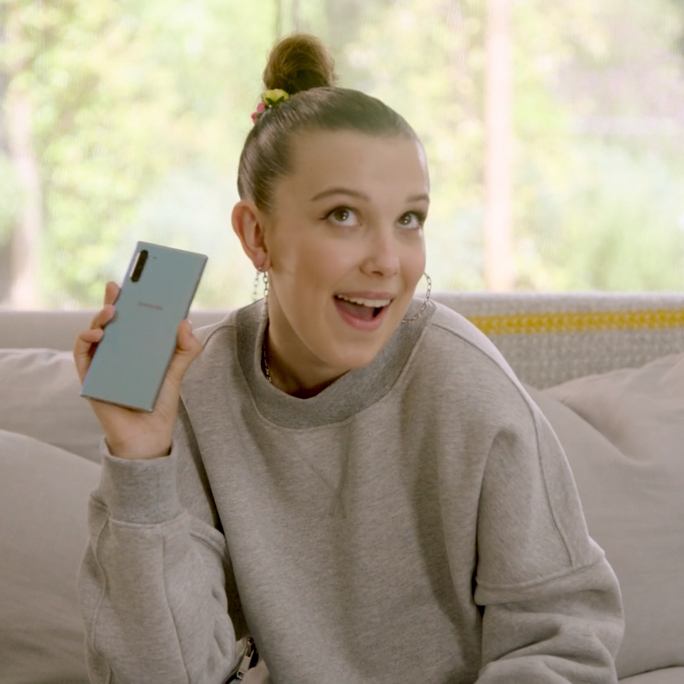 Video still of Millie Bobby Brown sitting on a couch, holding up an Aura Glow Galaxy Note10