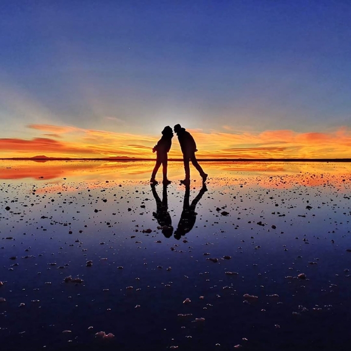 An image of a couple standing on a reflected surface with a  colorful sunset in the background, they are in playful poses facing eachother