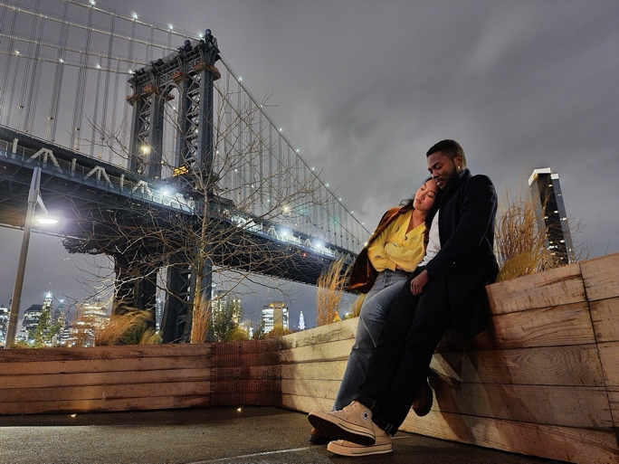 A couple sitting on a wooden surface with the Manhattan Bridge in the background, the scene is shot from a low angle