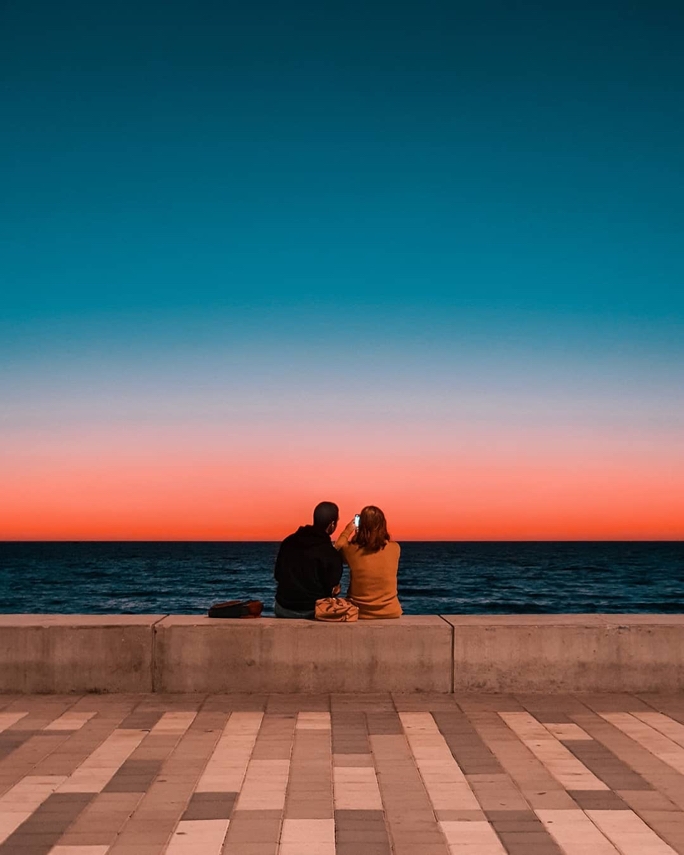 A couple sitting on a concrete wall looking out on the ocean, there is  a striking sunset that creates a colourful gradient in the sky.