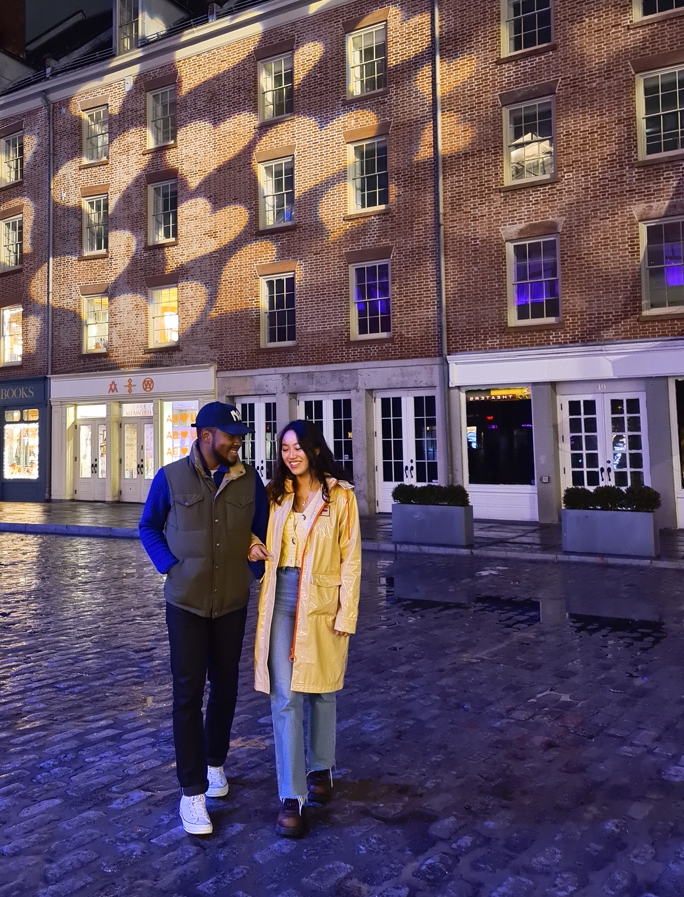 A smiling couple with linked arms walking down a cobbled street, there are heart-shaped lights being  projected onto the buildings in the background