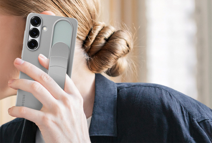 A woman holds a Galaxy S25+ installed with the Standing Grip Case in Gray. One finger holding the phone is tucked under the adjustable grip.