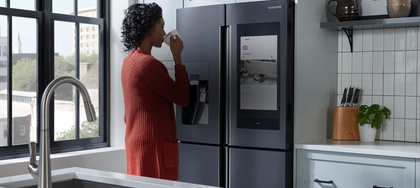 A young woman in her kitchen drinks from a mug while watching the screen of her smart refrigerator.