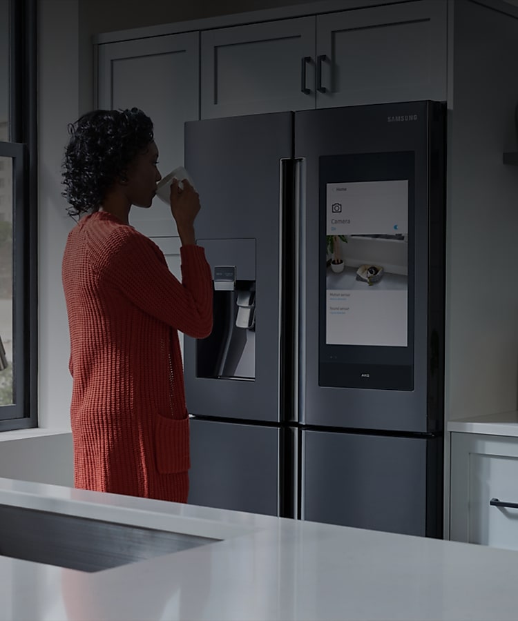 A young woman in her kitchen drinks from a mug while watching the screen of her smart refrigerator.