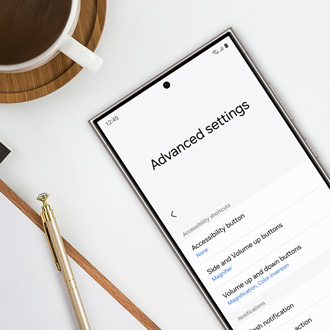 Galaxy phone on a desk displaying the 'Advanced settings' menu, with options for accessibility features, next to a notepad, pen, and a cup of coffe