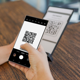 Hand holding a Galaxy phone scanning a QR code displayed on another device's screen in a well-lit office setting