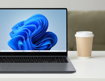 Samsung Galaxy Book laptop on a desk displaying a blue abstract wallpaper, with a disposable coffee cup beside it.