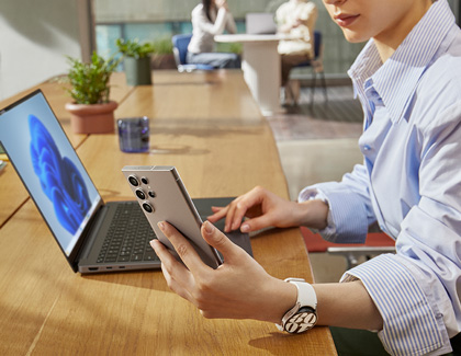 A woman in a business setting using a Galaxy phone to perform a software update via Smart Switch, with a laptop open on the table in front of her.