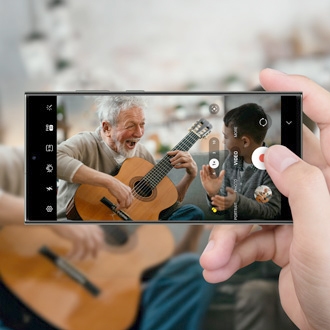 Hands holding a Galaxy phone recording an 8K video of two people playing guitars and laughing, displayed on the phone's camera interface.