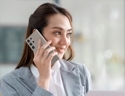 A young woman in a gray suit smiling while talking on a Galaxy phone, holding it to her ear. She appears to be in a bright, modern office environment.