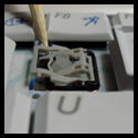 Macro shot of a scissor mechanism on a laptop keyboard, shown with a tool adjusting or fixing the alignment before replacing a keycap.