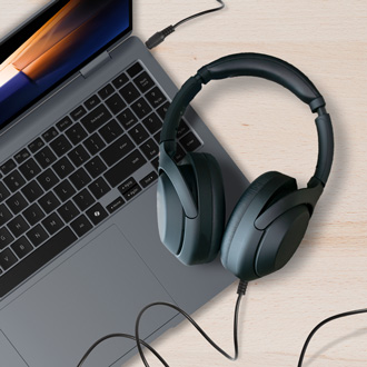 A pair of wired headphones resting next to a Samsung laptop on a wooden desk, with the headphone jack visibly connected to the laptop.