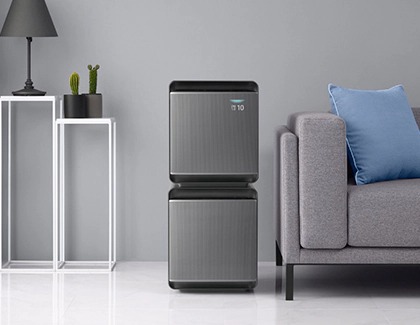 Two Samsung air purifiers stacked on top of each other next to a gray sofa in a modern living room.