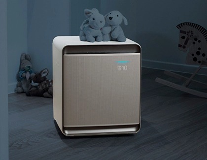 A Samsung air purifier placed in a dimly lit children's room with stuffed animals nearby.