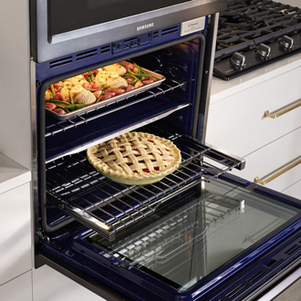 An open Samsung oven with a Flex Duo feature, showing a baking tray with various dishes on the upper rack and a pie on the lower rack.