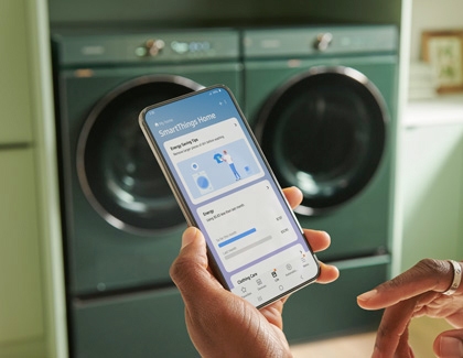 A person holding a Galaxy phone with the SmartThings app open in front of a Samsung washer and dryer
