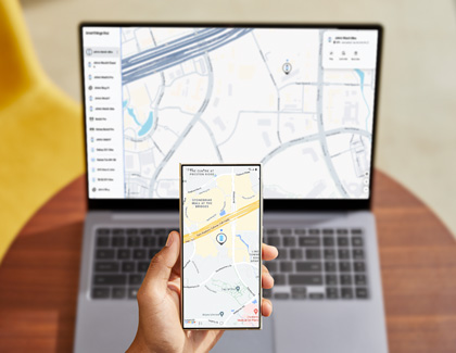 A hand holding a Galaxy phone while displaying the location of a Galaxy device whilst in front of a Galaxy Book displaying the SmartThings Find website. 