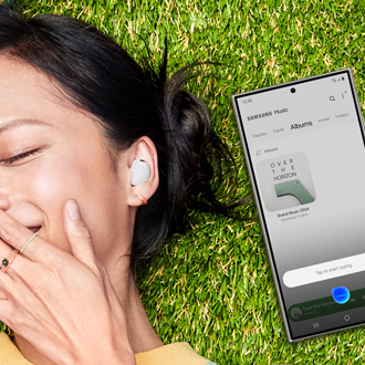 A woman lying on grass with Galaxy Buds2 Pro in her ears, next to a Galaxy S24 Ultra showing Bixby