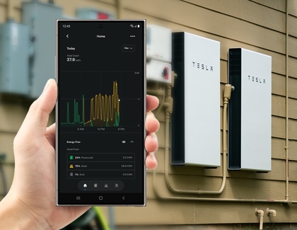 A hand holding a Galaxy phone showing energy consumption data, with Tesla Powerwall units installed on the wall in the background.