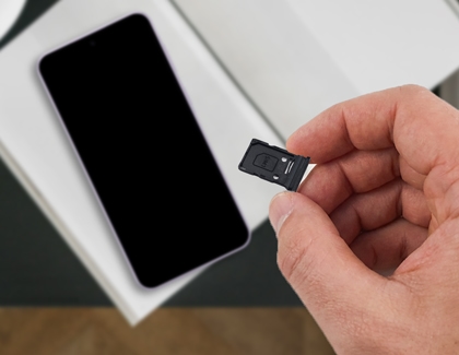 A hand holding a black SIM card tray with a SIM card inside next to a purple Galaxy A series phone.