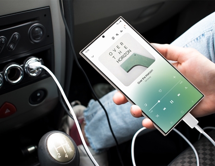 Hand holding a Samsung Galaxy phone connected to a car via USB, showing a music player interface.
