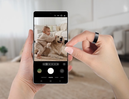 Person using hand gestures with the Galaxy Ring to control the Galaxy S25 Ultra's camera, capturing a photo of a baby playing with a dog.