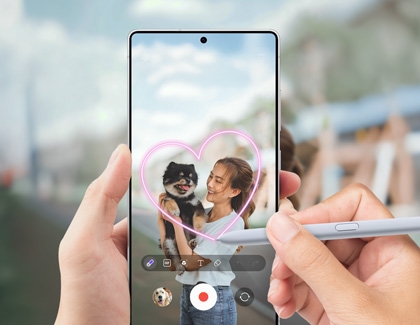 A person using a Galaxy S25 Ultra while drawing a pink heart with an S Pen using the AR Doodle feature while capturing a photo of a woman holding a dog