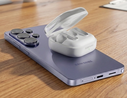A white Galaxy Buds4 Pro case charging wirelessly on the back of a purple Galaxy S26 Ultra on a wooden table.