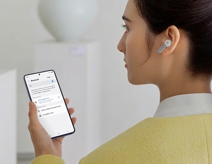 A woman looks at a Galaxy S26 Ultra showing Bluetooth settings while wearing a Galaxy Buds4 Pro.