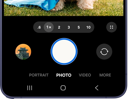 A Galaxy phone showing the Camera app in Photo mode with the shutter button highlighted in blue and zoom options displayed.