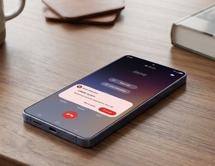 A Galaxy phone lying on a wooden table showing an incoming call screen with a scam detection warning popup displayed.
