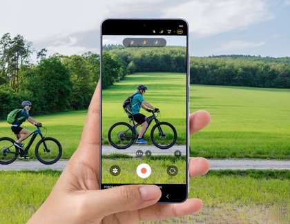 A hand holds a purple Galaxy S26 Ultra recording a person on a bicycle in a green field with Super steady mode options visible on the screen.