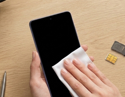 A hand wiping down a Galaxy phone with a cloth, with a removed SIM tray visible on the desk behind it.