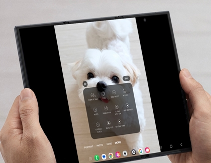 Hands holding the unfolded Galaxy Z TriFold showing the camera interface with a More menu overlaying a photo of a white dog.
