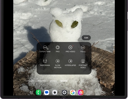 A Galaxy Z Fold showing the camera app with More selected, displaying camera modes over a snowman image on a tree stump.
