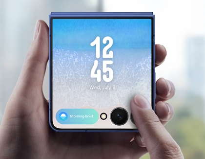 A hand holding a folded Galaxy Z Flip7 showing the FlexWindow with the time, date, and a Morning brief notification.