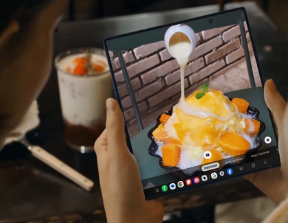 Person holding the Galaxy Z Fold7 unfolded, capturing a dessert with the Camera app while seated at a table in a dim cafe.