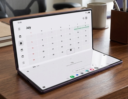 A Galaxy Z Fold device partially folded on a wooden desk showing a calendar app on its main display.