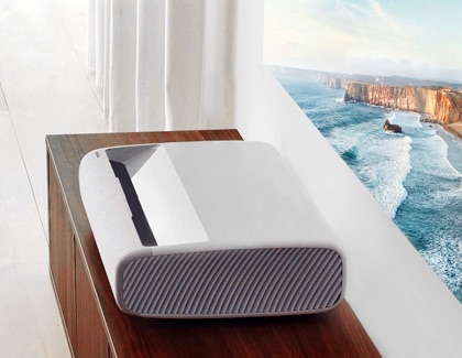 Samsung Premiere projector placed on a wooden cabinet projecting a coastal landscape onto a nearby wall.