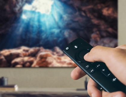 Hand holding an Amazon Fire Stick remote controlling a Samsung TV displaying a cave scene with sunlight streaming in.