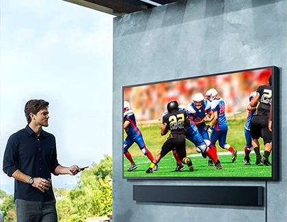 A man watching a football game on a mounted Samsung TV outdoors.