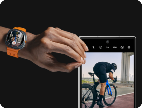 A hand wearing a Galaxy Watch Ultra is making a Double Pinch motion. Next to it is the paired Galaxy Z Fold6 with its Cover screen featuring a camera screen capturing a man cycling.
