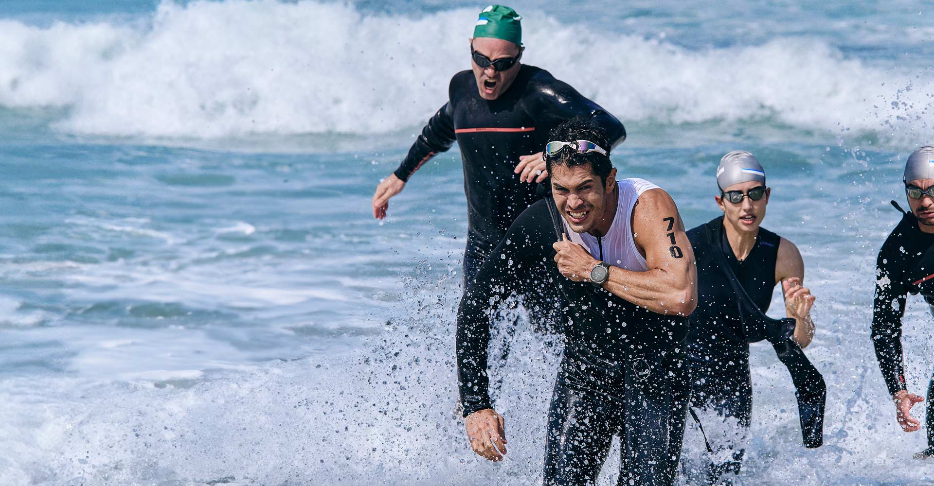 Triathlon athletes are running out of the ocean water after swimming. One man wearing a Galaxy Watch Ultra is taking off his swimsuit to transition into the next sport.